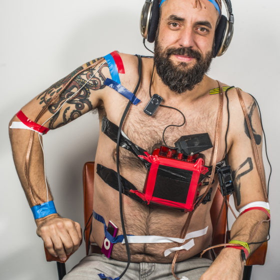 Jonny smiling, topless wearing headphones with sound equipment taped to his chest