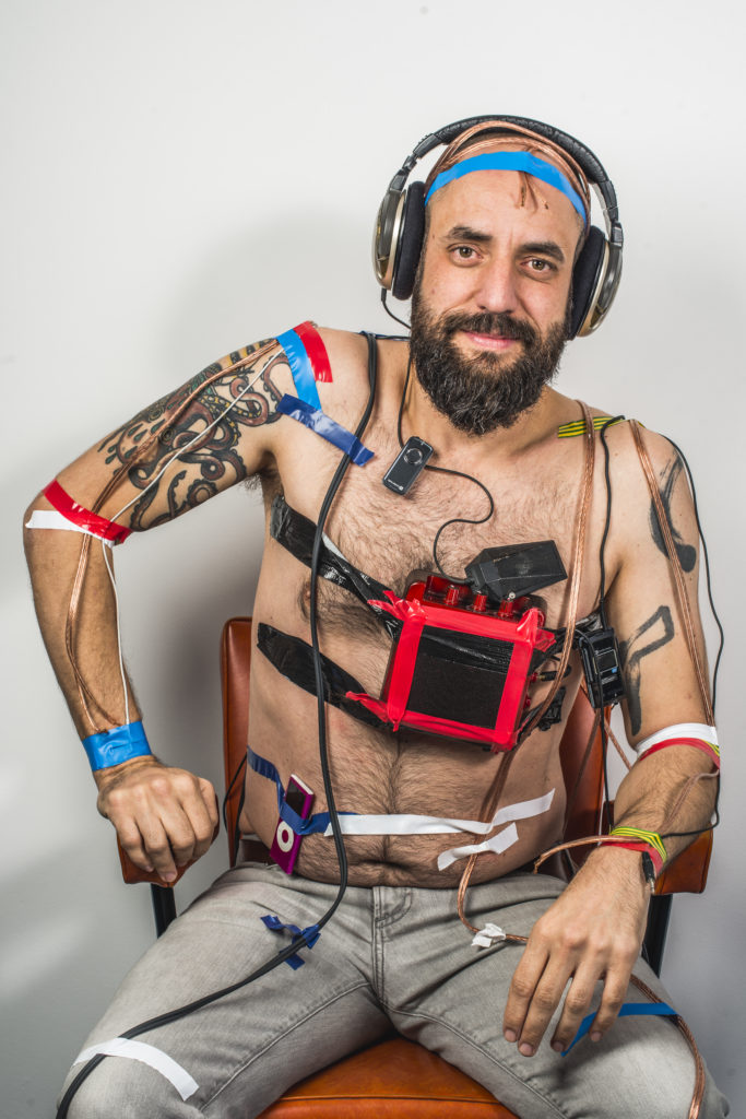 Jonny smiling, topless wearing headphones with sound equipment taped to his chest